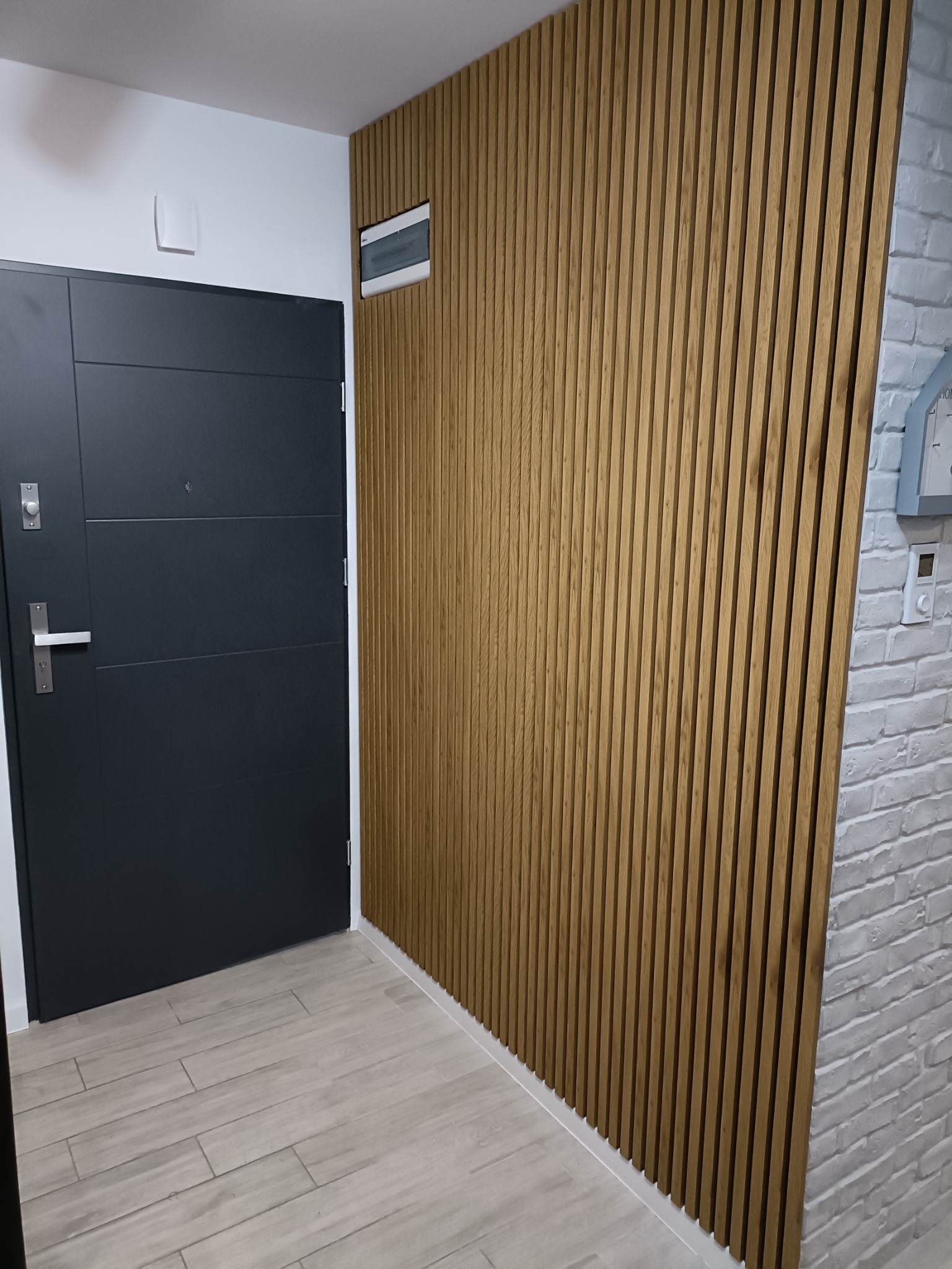 Nowoczesny przedpokój z drewnianą ścianą z lameli, szara drzwi wejściowe i podłoga imitująca drewno, obok ściana z cegły pomalowana na biało.