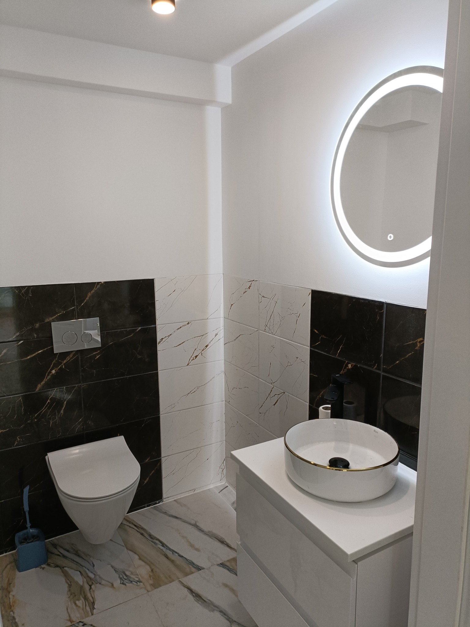 Nowoczesna łazienka z czarno-białymi płytkami imitującymi marmur, podwieszana toaleta, umywalka nablatowa z złotym rantem i okrągłe lustro z podświetleniem LED.