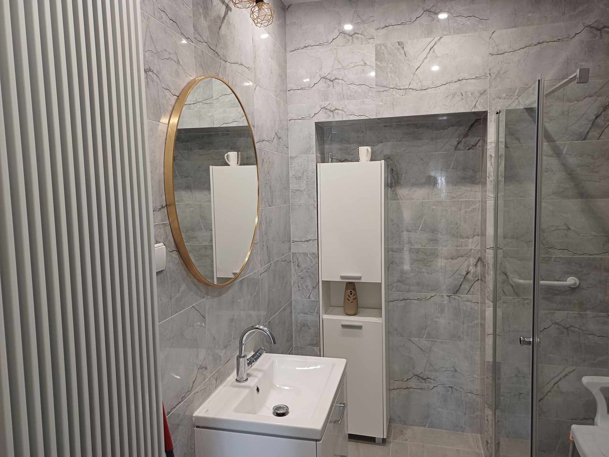 Nowoczesna łazienka z szarymi marmurkowymi płytkami, okrągłym lustrem w złotej ramie, białą szafką i kabiną prysznicową ze szklanymi drzwiami.