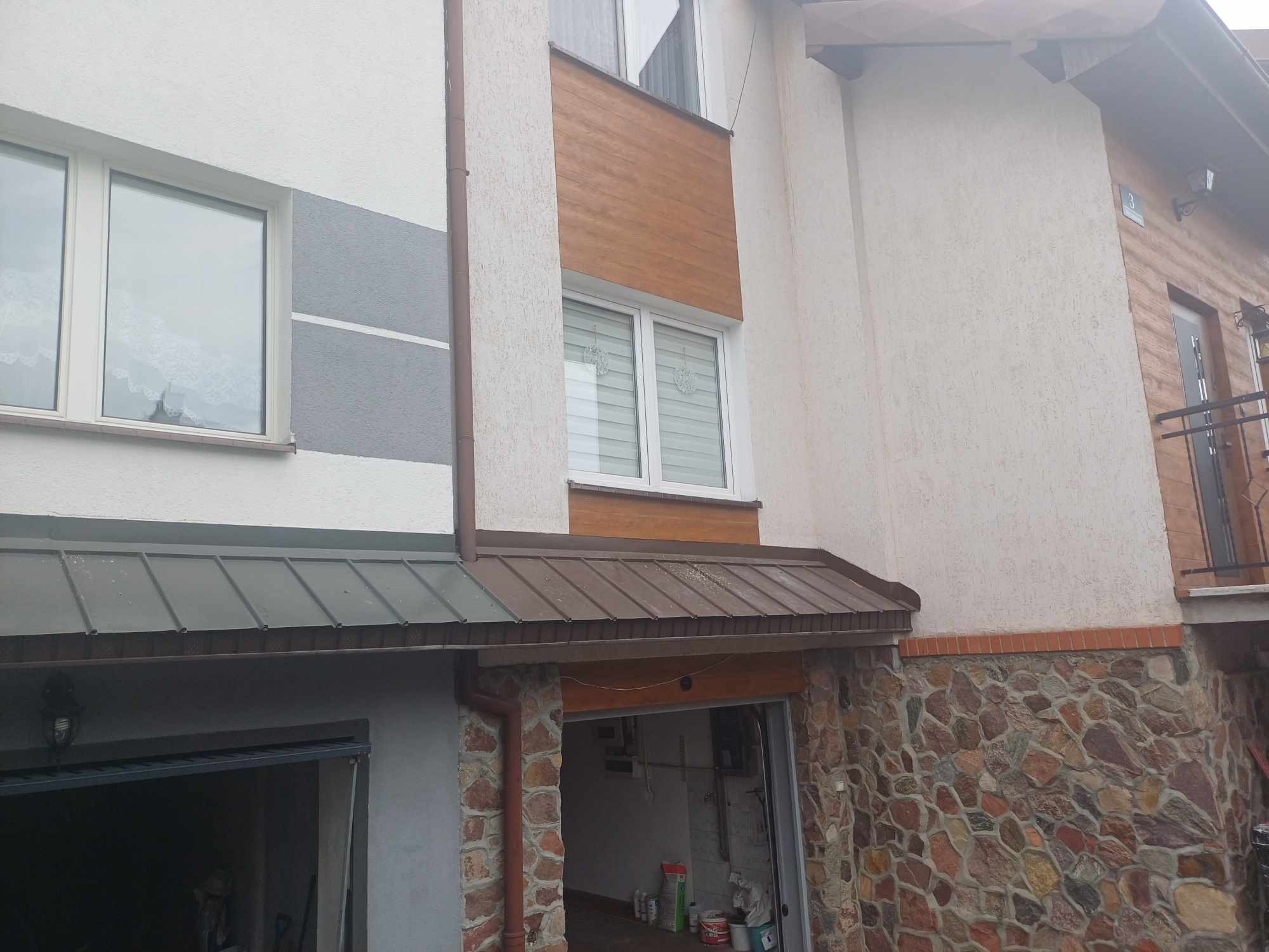 Zewnętrzny widok dwupiętrowego domu z garażem i kamienną elewacją, z różnymi fakturami tynku i brązowym dachem nad garażem.