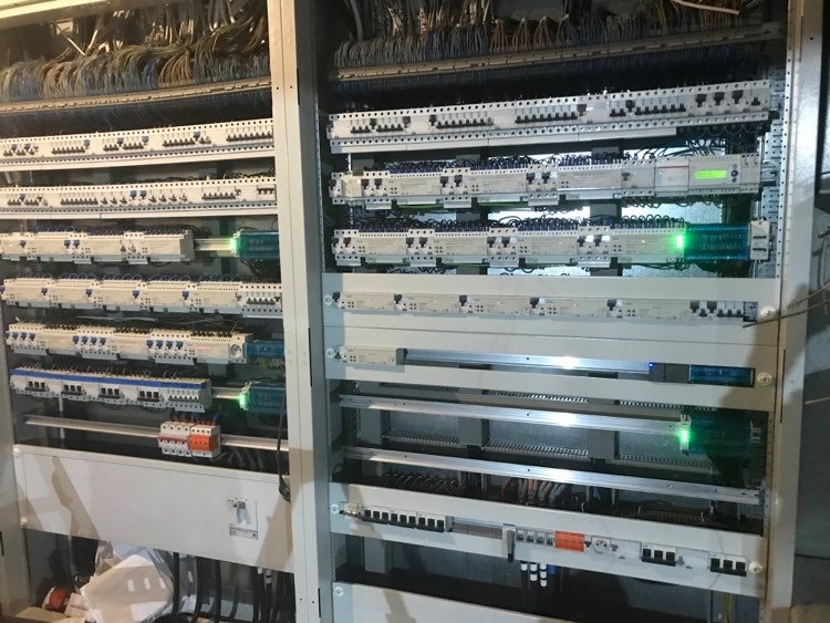 Rozdzielnica elektryczna z licznymi modułami, przewodami i zielonymi diodami sygnalizacyjnymi w szarej obudowie.