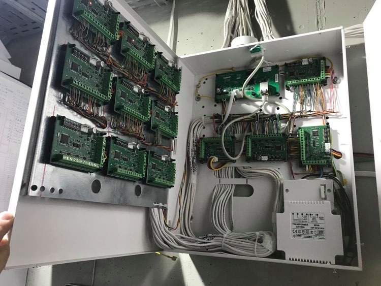 Otwarta biała skrzynka rozdzielcza z licznymi zielonymi płytkami drukowanymi i kablami, widoczny transformator NOVA AWT500 na dole.