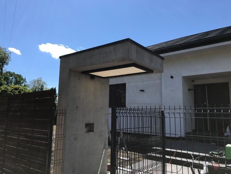 Betonowy słup bramy z wbudowanym oświetleniem LED, widok z zewnątrz posesji przez metalowe ogrodzenie panelowe.