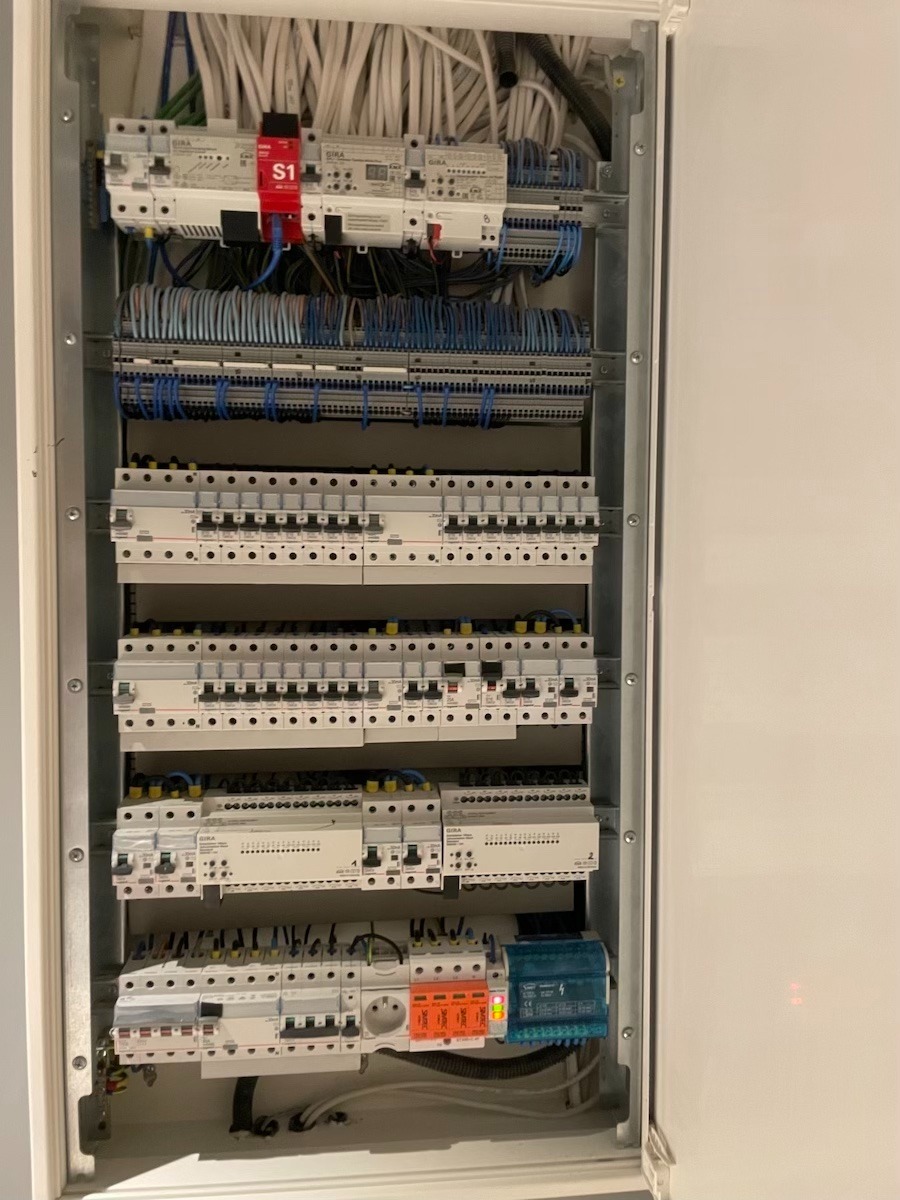 Otwarta skrzynka elektryczna z licznymi bezpiecznikami, przewodami i modułami, widoczne marki GIRA, jeden z modułów oznaczony czerwonym paskiem S1.