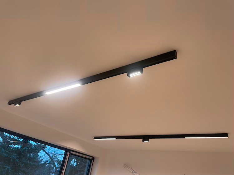 Dwie czarne szyny z oświetleniem LED zamontowane na białym suficie, widok z dołu, okno z widokiem na drzewa w tle.