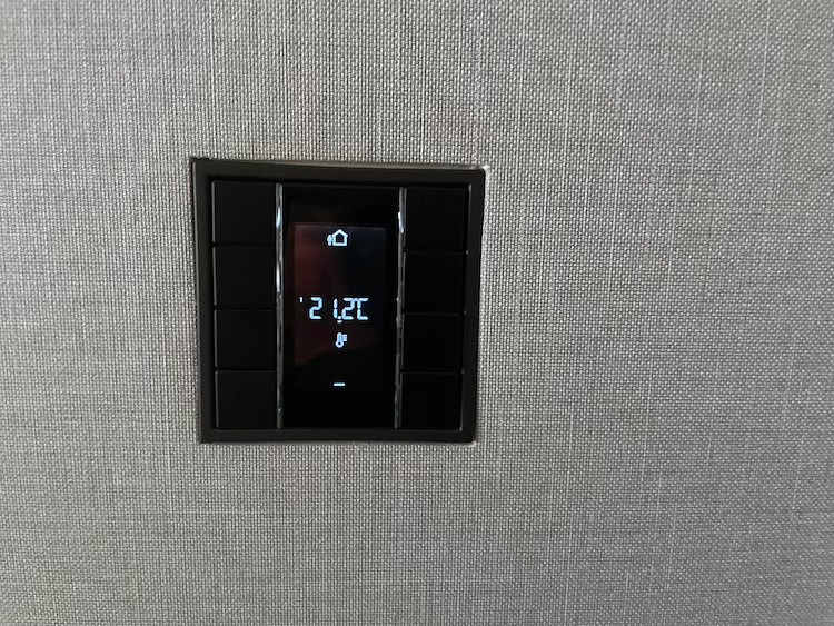 Czarny panel sterowania inteligentnego domu, wpuszczony w szarą ścianę o teksturze tkaniny, wyświetla temperaturę 21.2 stopni Celsjusza oraz ikonę domu.