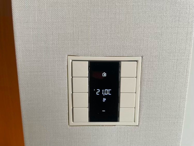 Panel sterowania inteligentnego domu z wyświetlaczem temperatury ustawionej na 21 stopni Celsjusza, wbudowany w ścianę o teksturowanej powierzchni.