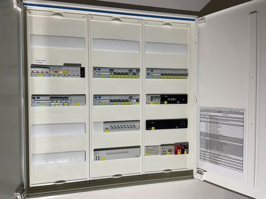 Otwarta, rozbudowana szafa elektryczna z widocznymi zabezpieczeniami, modułami sterującymi i schematem instalacji na wewnętrznej stronie drzwi.
