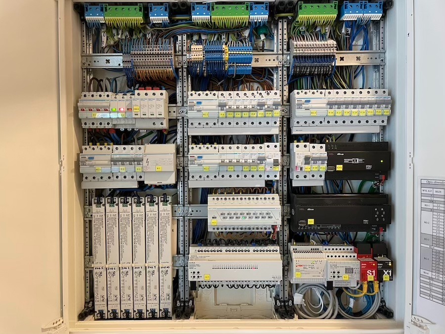 Rozdzielnica elektryczna z licznymi zabezpieczeniami, przewodami i elementami sterującymi, widoczne oznaczenia obwodów i etykiety identyfikacyjne.