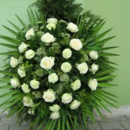 Okrągły wieniec z białych róż i goździków, otoczony długimi, zielonymi liśćmi, na tle zielonej ściany i szarej kostki brukowej.