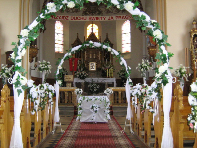 Udekorowane wnętrze kościoła na uroczystość ślubną: białe kwiaty i tiul zdobią ławki, ołtarz i tworzą ozdobne łuki.