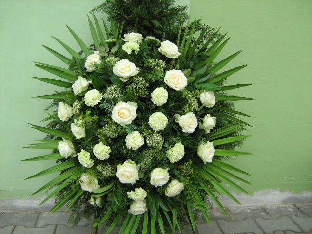 Okrągły wieniec z białych róż i goździków, otoczony długimi, zielonymi liśćmi, na tle zielonej ściany i szarej kostki brukowej.