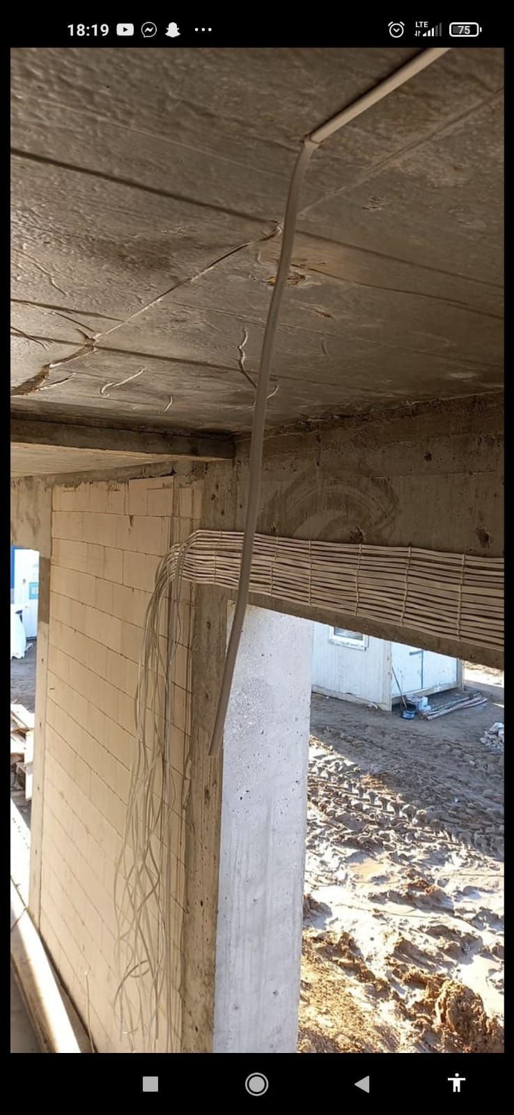 Kable elektryczne prowadzone wzdłuż surowego betonowego sufitu i ściany budynku w trakcie budowy, widoczne korytka kablowe, tło stanowi plac budowy z barakiem.