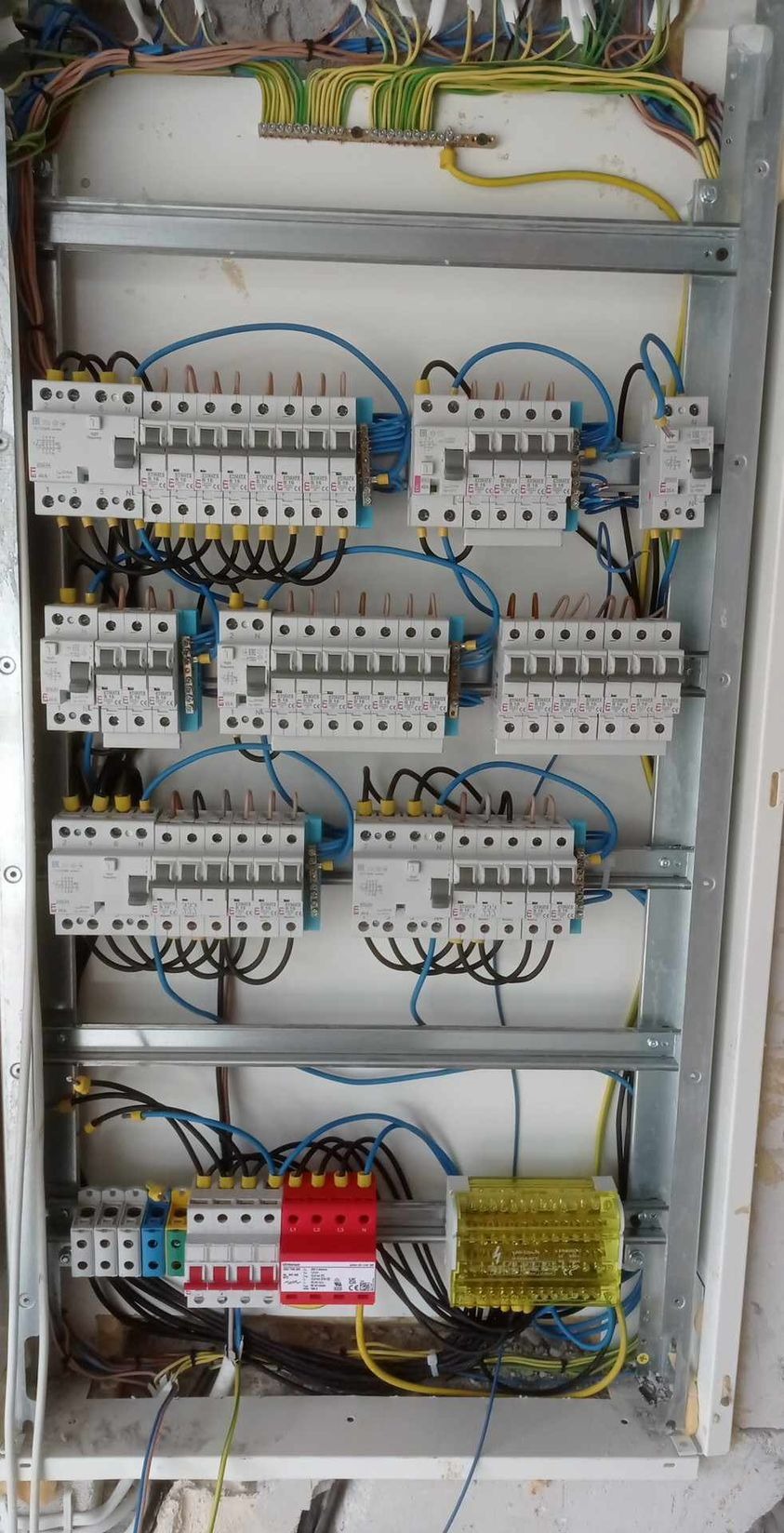 Rozdzielnica elektryczna z zamontowanymi bezpiecznikami, widoczne przewody w kolorach niebieskim, czarnym, żółto-zielonym, oraz czerwony ogranicznik przepięć. Metalowa obudowa rozdzielnicy...