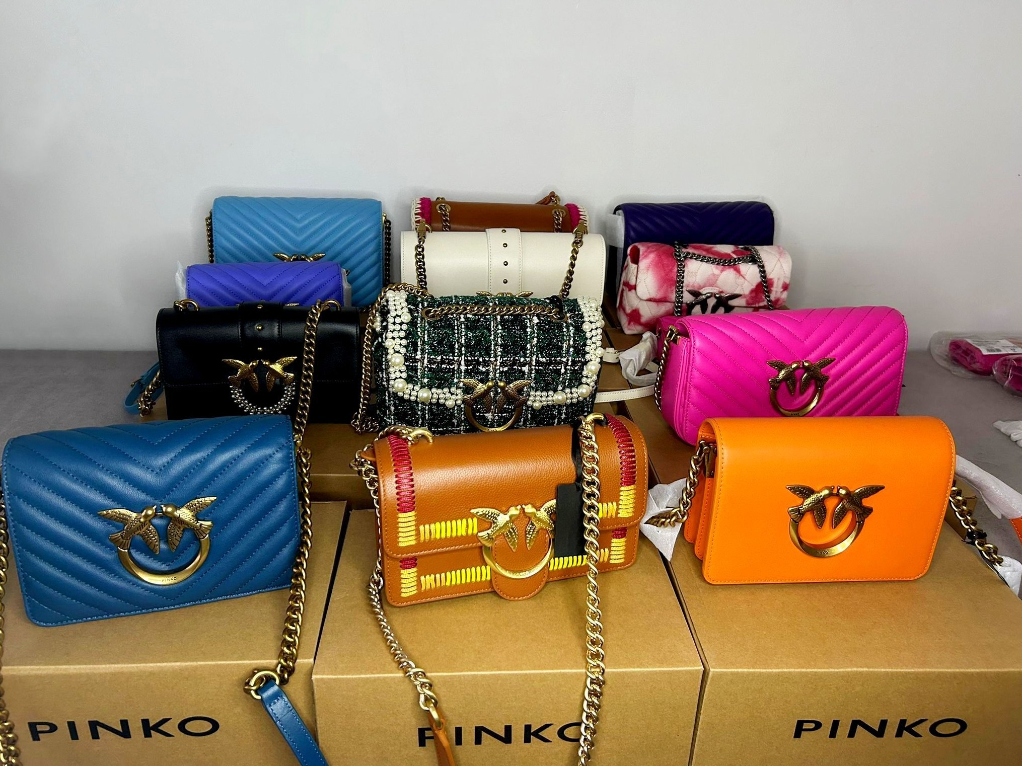 Kolekcja torebek marki Pinko w różnych kolorach i wzorach, z charakterystycznym logo dwóch ptaków, ułożonych na kartonowych pudełkach z napisem PINKO.