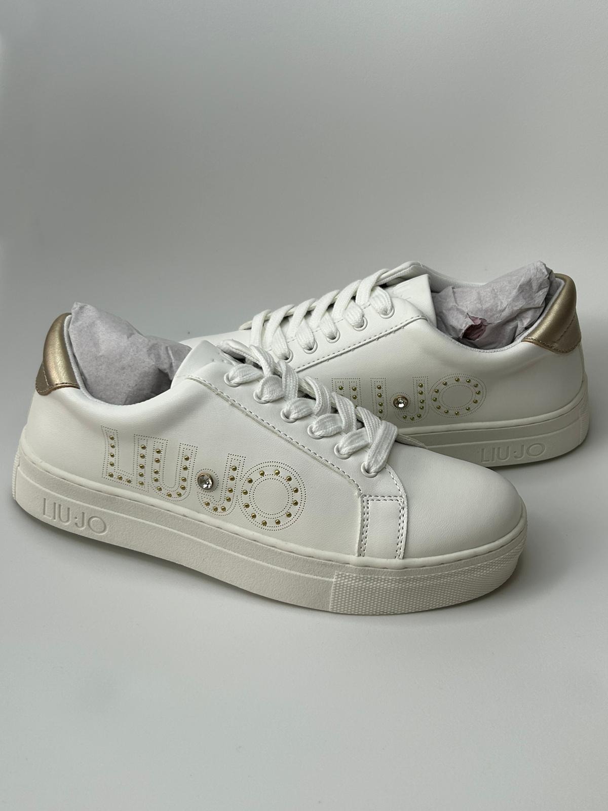 Dwa białe skórzane sneakersy marki Liu Jo z złotymi akcentami na pięcie i logo zdobionym złotymi ćwiekami i kryształkiem, na białym tle.