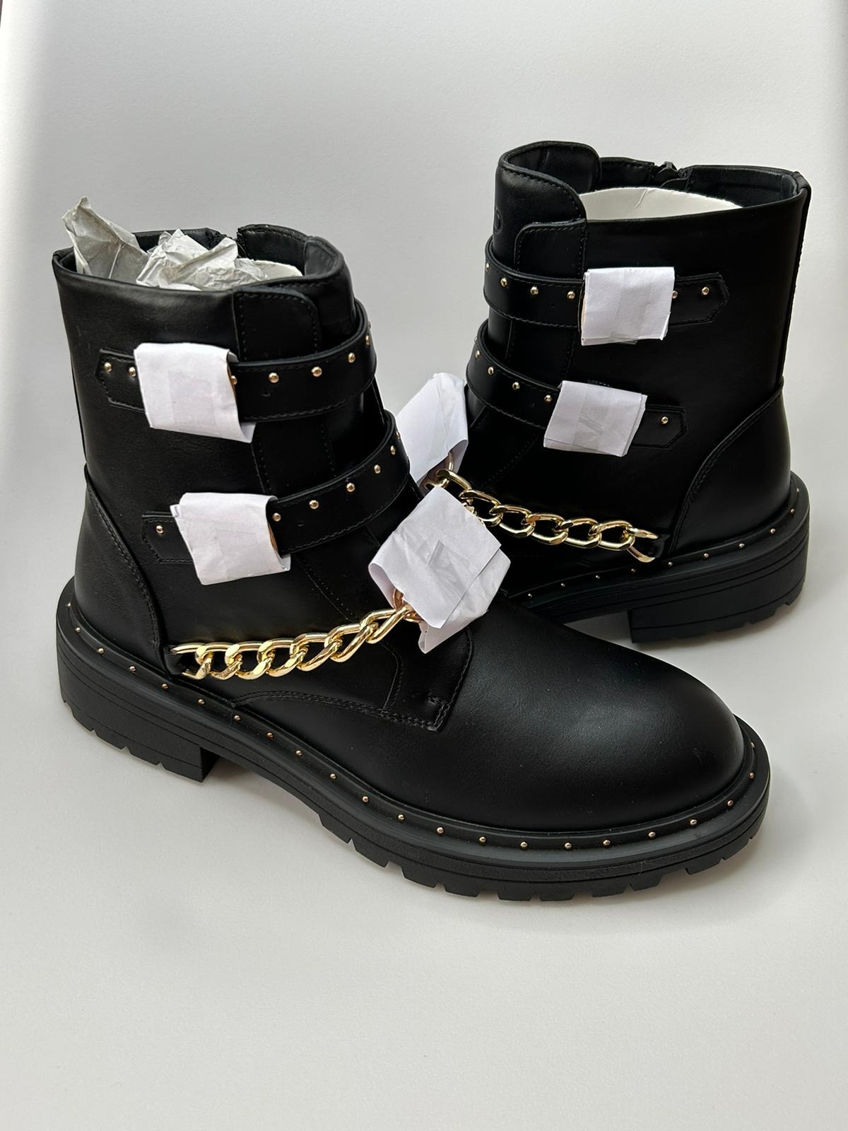 Para czarnych botków motocyklowych z złotymi łańcuchami i ćwiekami, zabezpieczonych białym papierem, na białym tle.