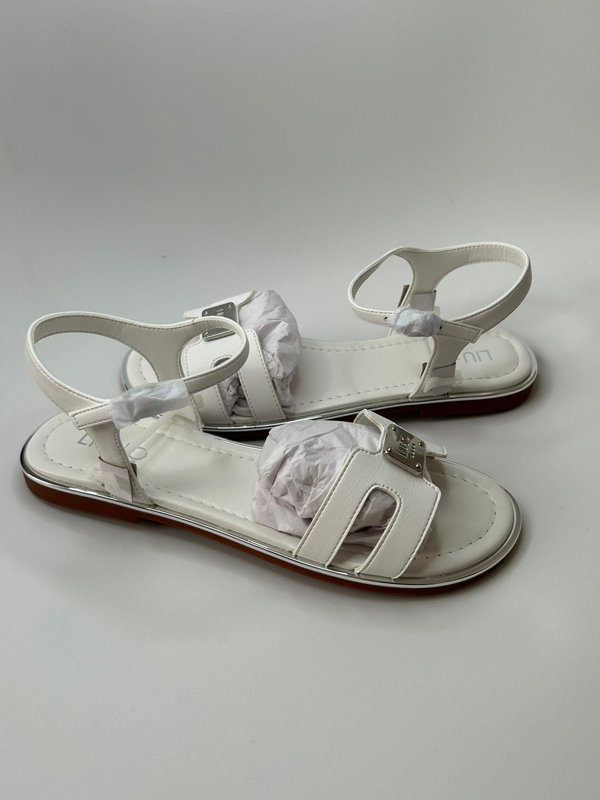 Para białych skórzanych sandałów marki Liu Jo z srebrnymi detalami i częściowo wypełnionych papierem, na białym tle.