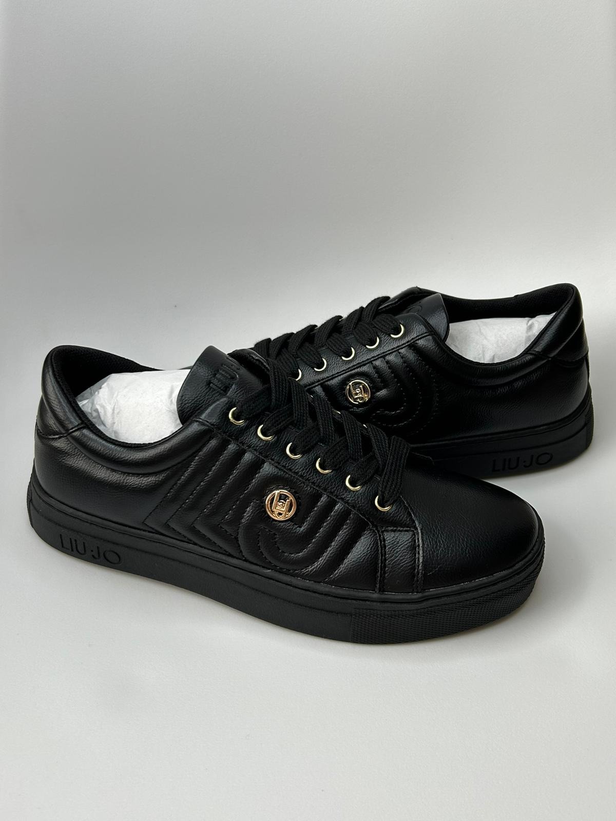 Para czarnych, skórzanych sneakersów marki Liu Jo z pikowanym wzorem i złotym logo na boku, sznurowane czarnymi sznurówkami, na jasnym tle.