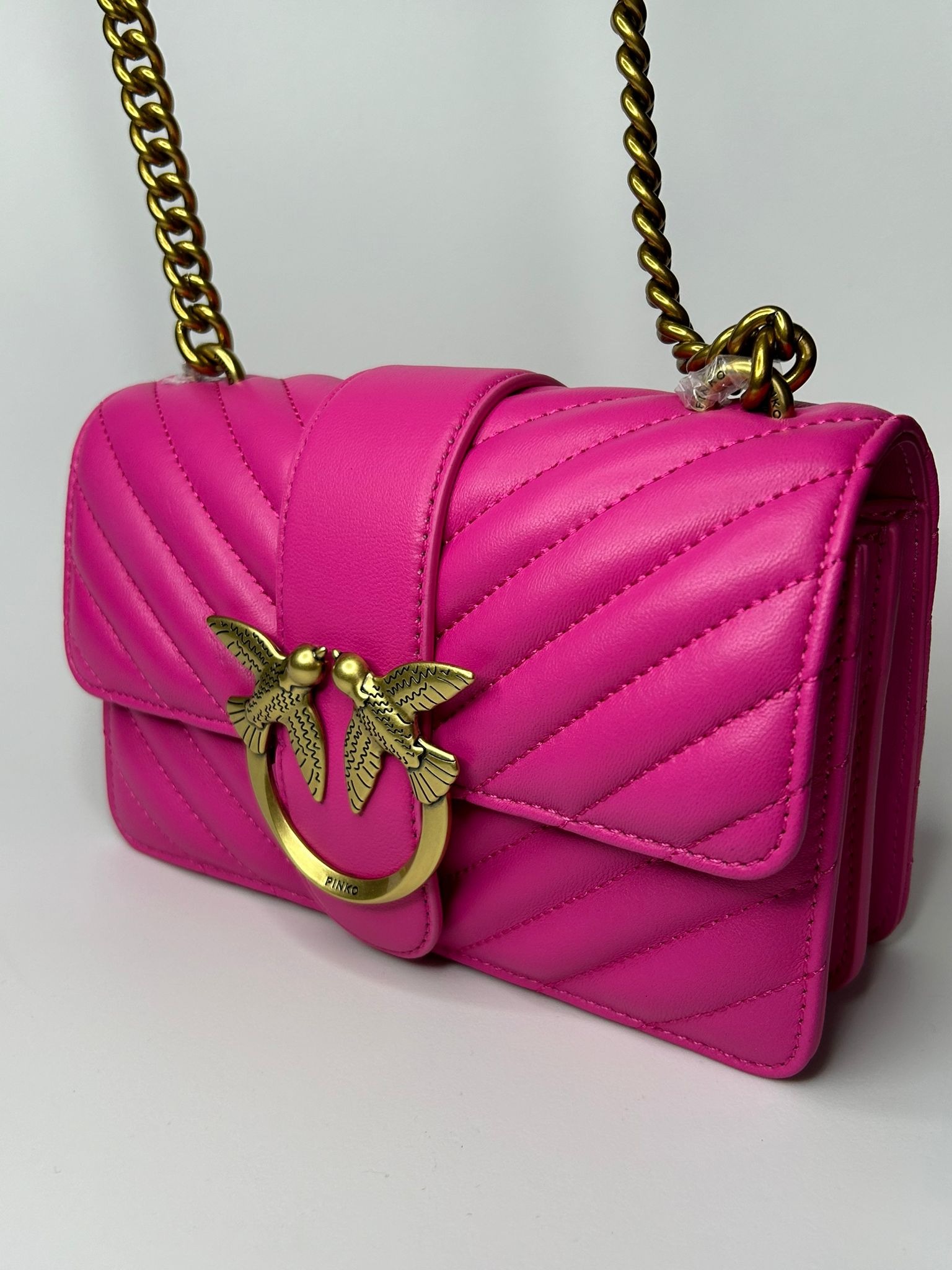 Fuksjowa, pikowana torebka skórzana na złotym łańcuszku, z metalowym logo dwóch ptaków stykających się dziobami, na jasnym tle.