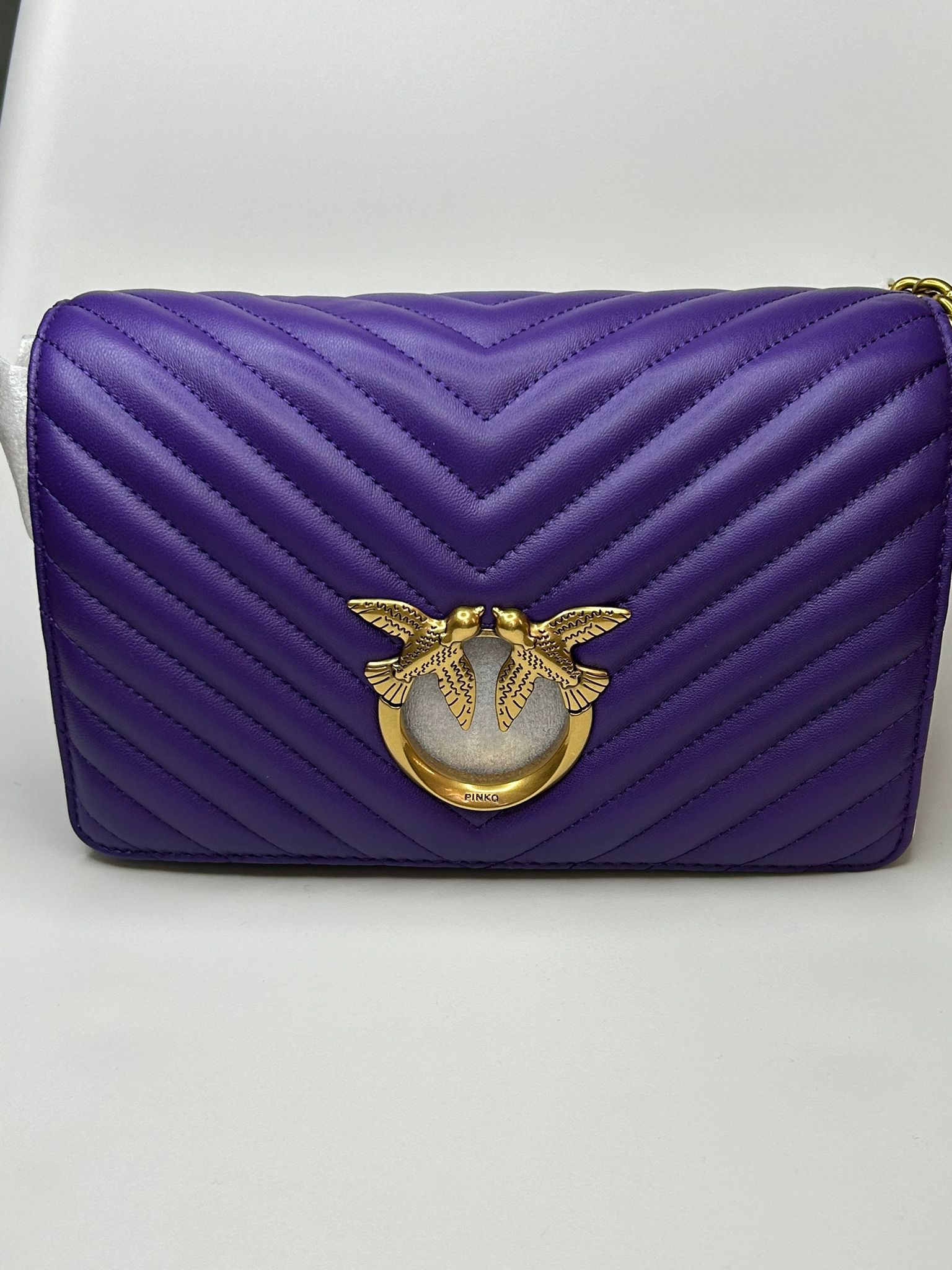 Fioletowa skórzana torebka z pikowaniem w jodełkę i złotym logo dwóch ptaków na białym tle.