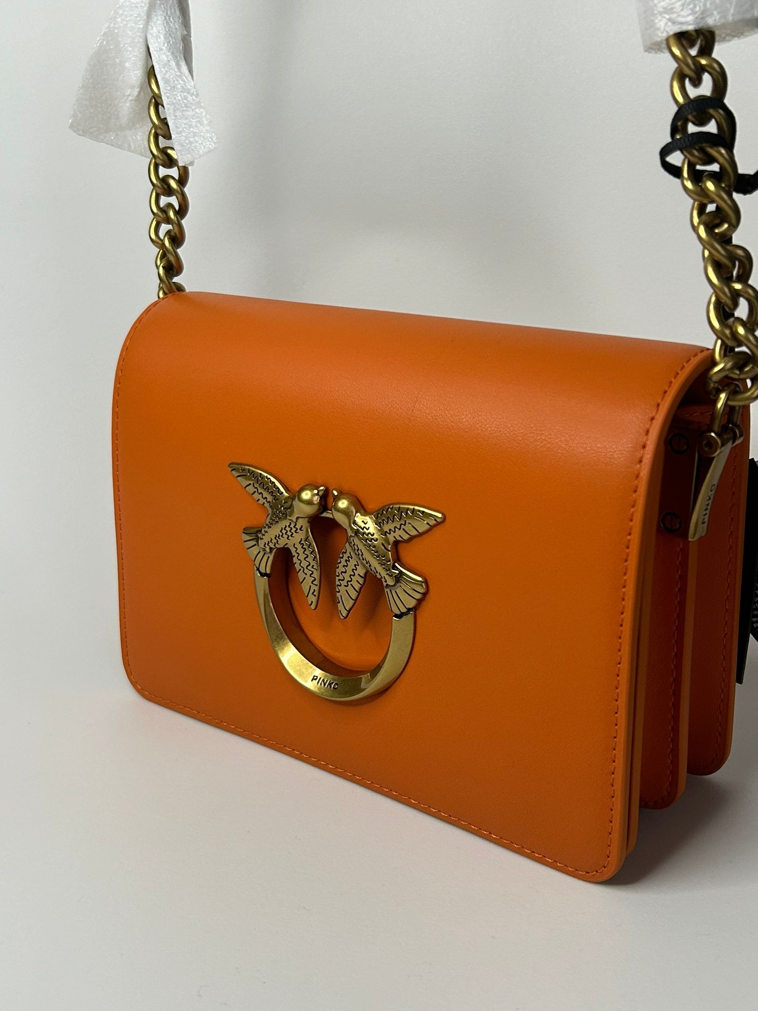 Pomarańczowa skórzana torebka listonoszka Pinko z złotym łańcuszkiem i metalowym logo z dwoma ptakami.