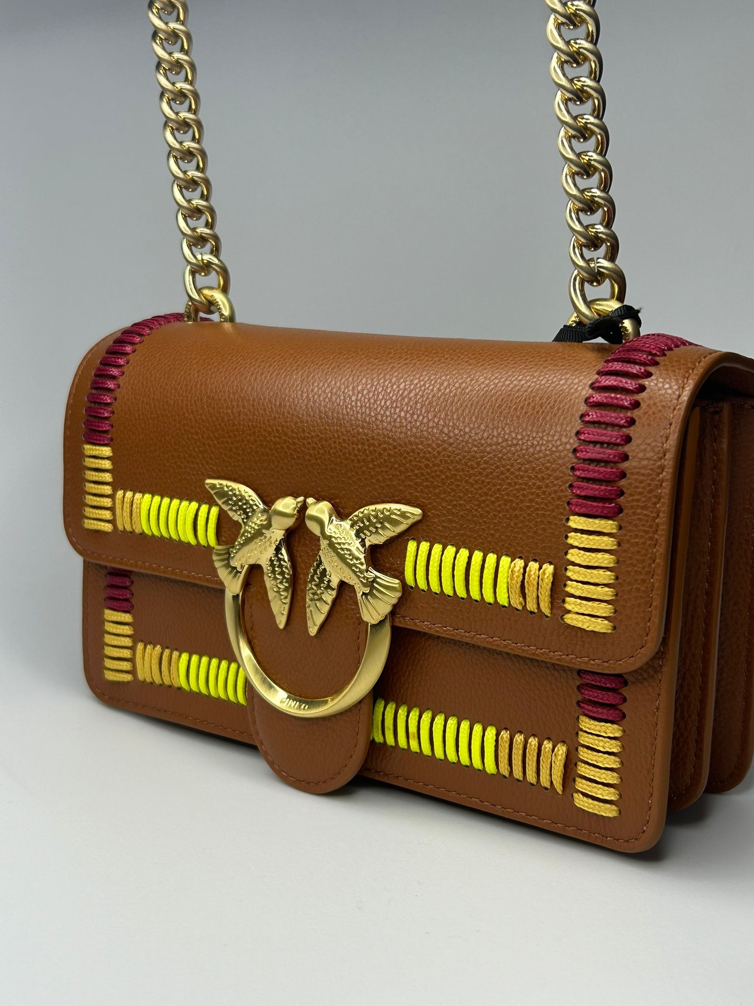 Brązowa skórzana torebka listonoszka z złotym łańcuszkiem i aplikacją z dwoma ptakami, zdobiona neonowo-żółtymi i bordowymi przeszyciami.