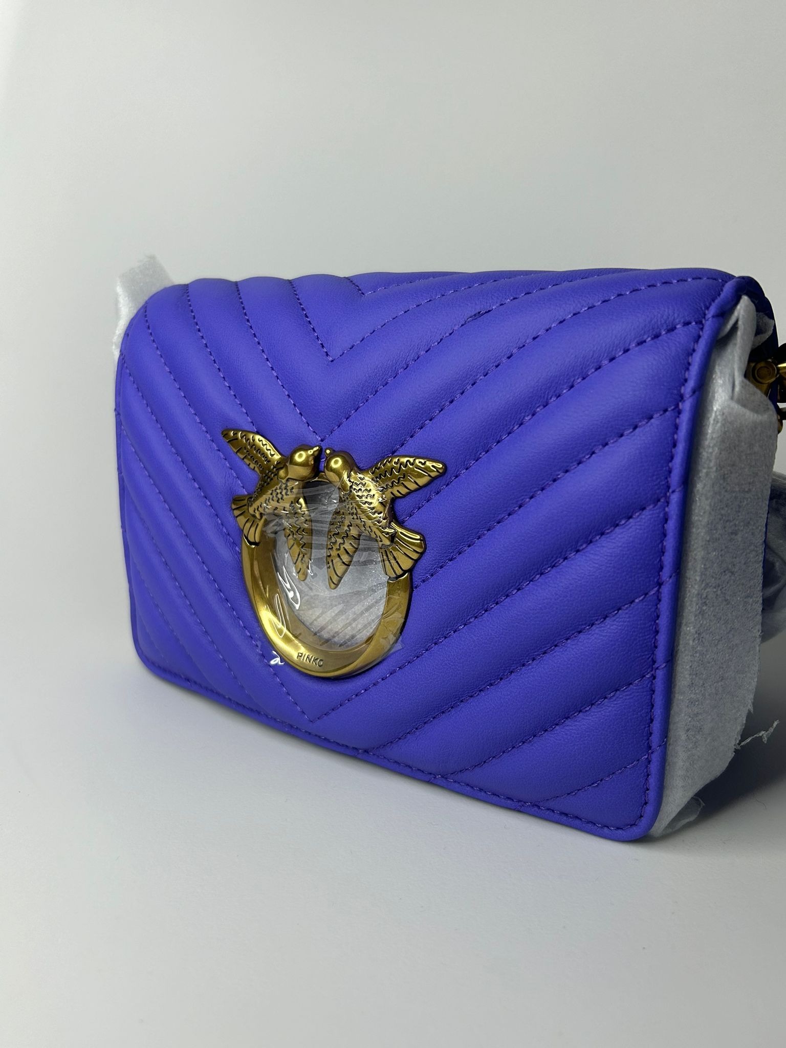Fioletowa skórzana torebka Pinko z pikowanym wzorem chevron, złotym logo z ptakami i folią ochronną na metalowych elementach, na białym tle.