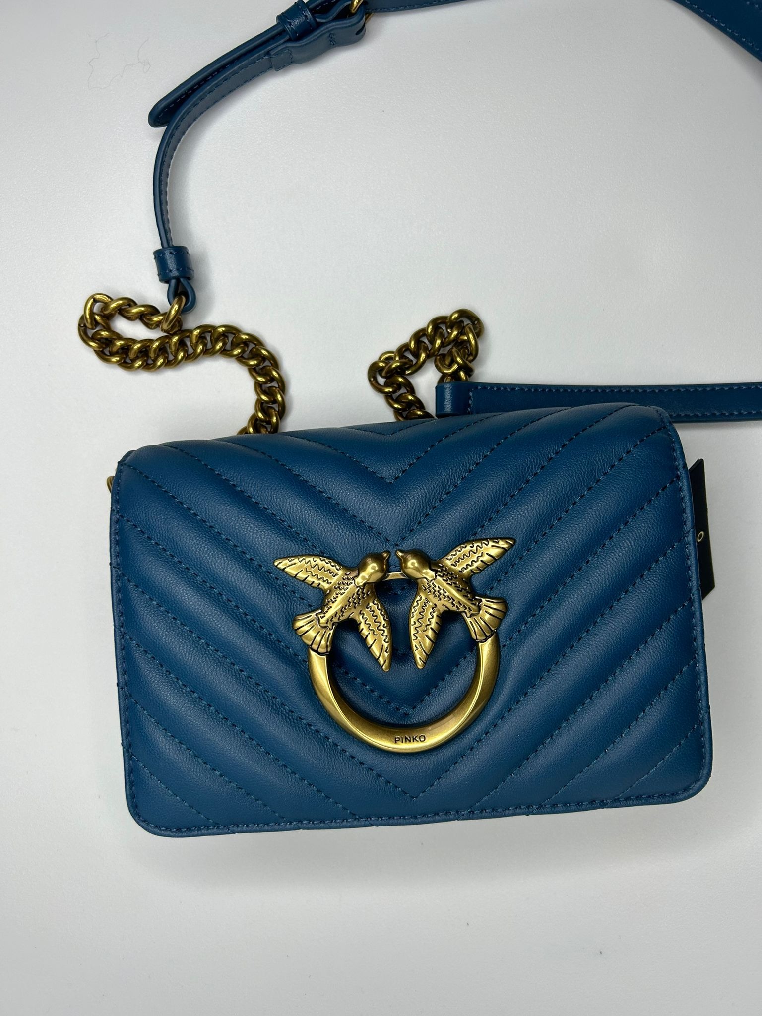 Turkusowa, pikowana torebka damska z metalowym logo dwóch ptaków i złotym łańcuszkiem na białym tle.