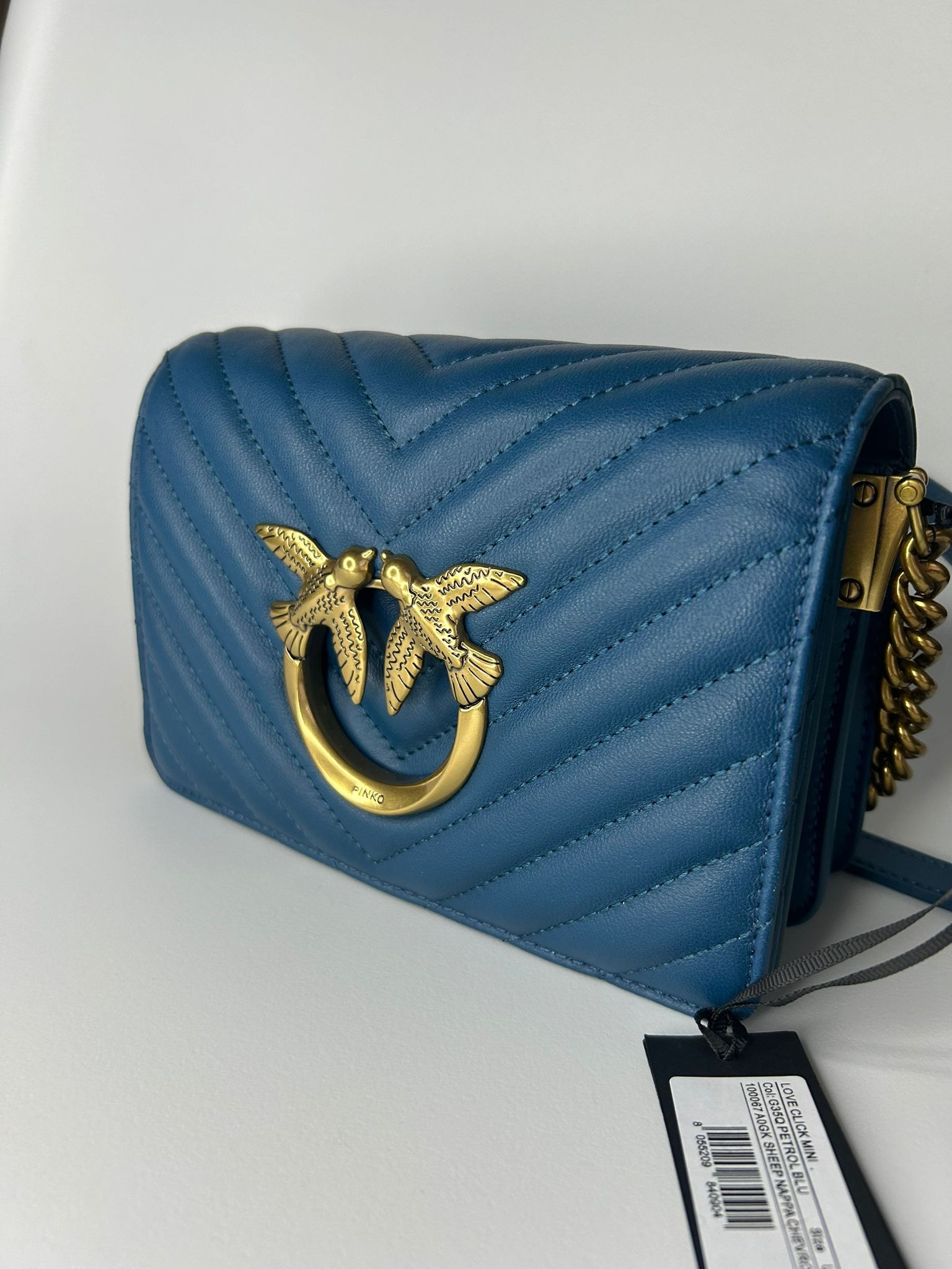 Niebieska skórzana torebka Pinko z charakterystycznym złotym logo dwóch całujących się ptaków na tle pikowanego wzoru, widoczna metka produktowa.