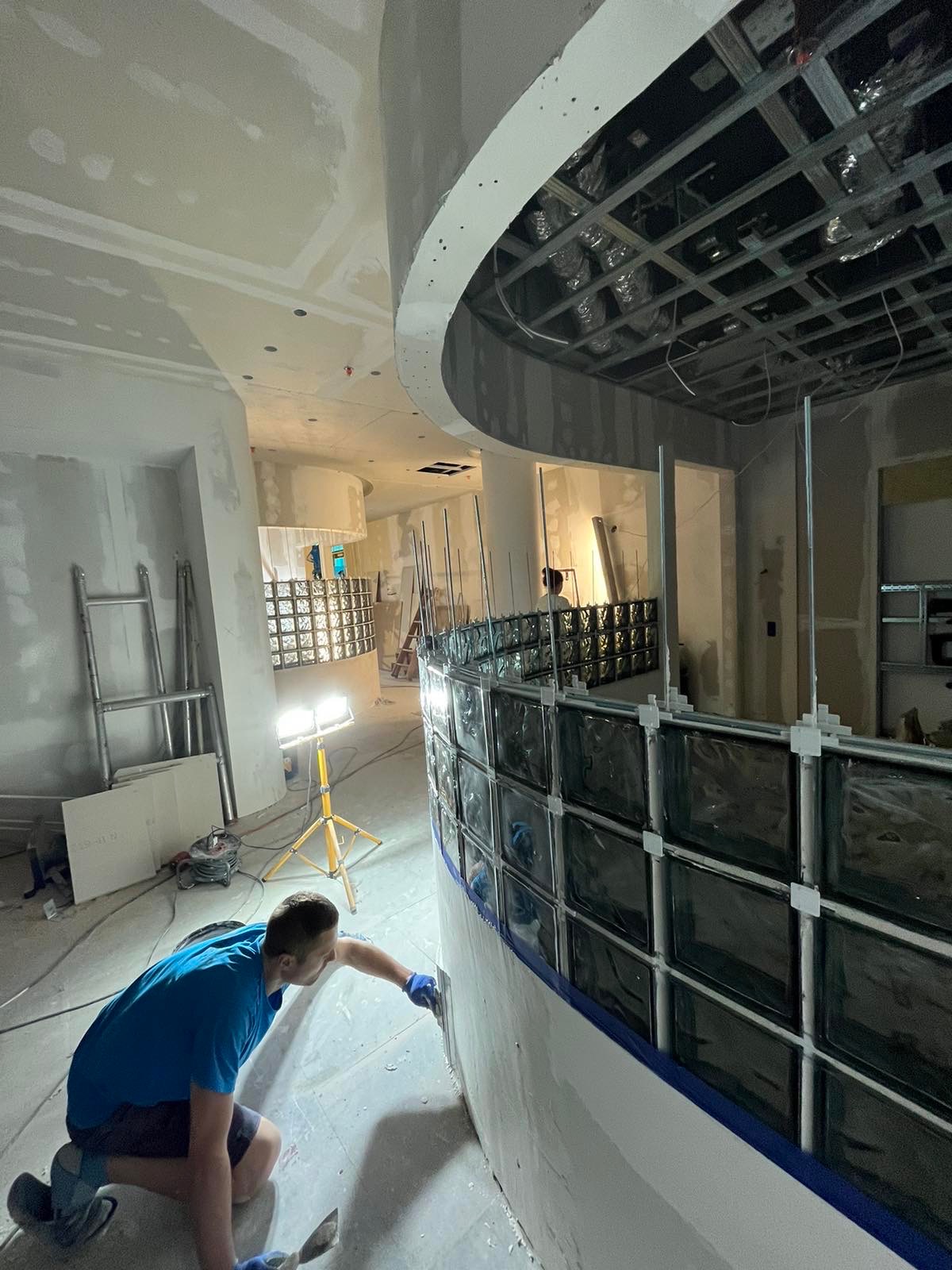 Pracownik szpachluje zakrzywioną ścianę z luksferów w surowym wnętrzu biura, widoczne metalowe profile sufitowe, lampa budowlana i drabina.