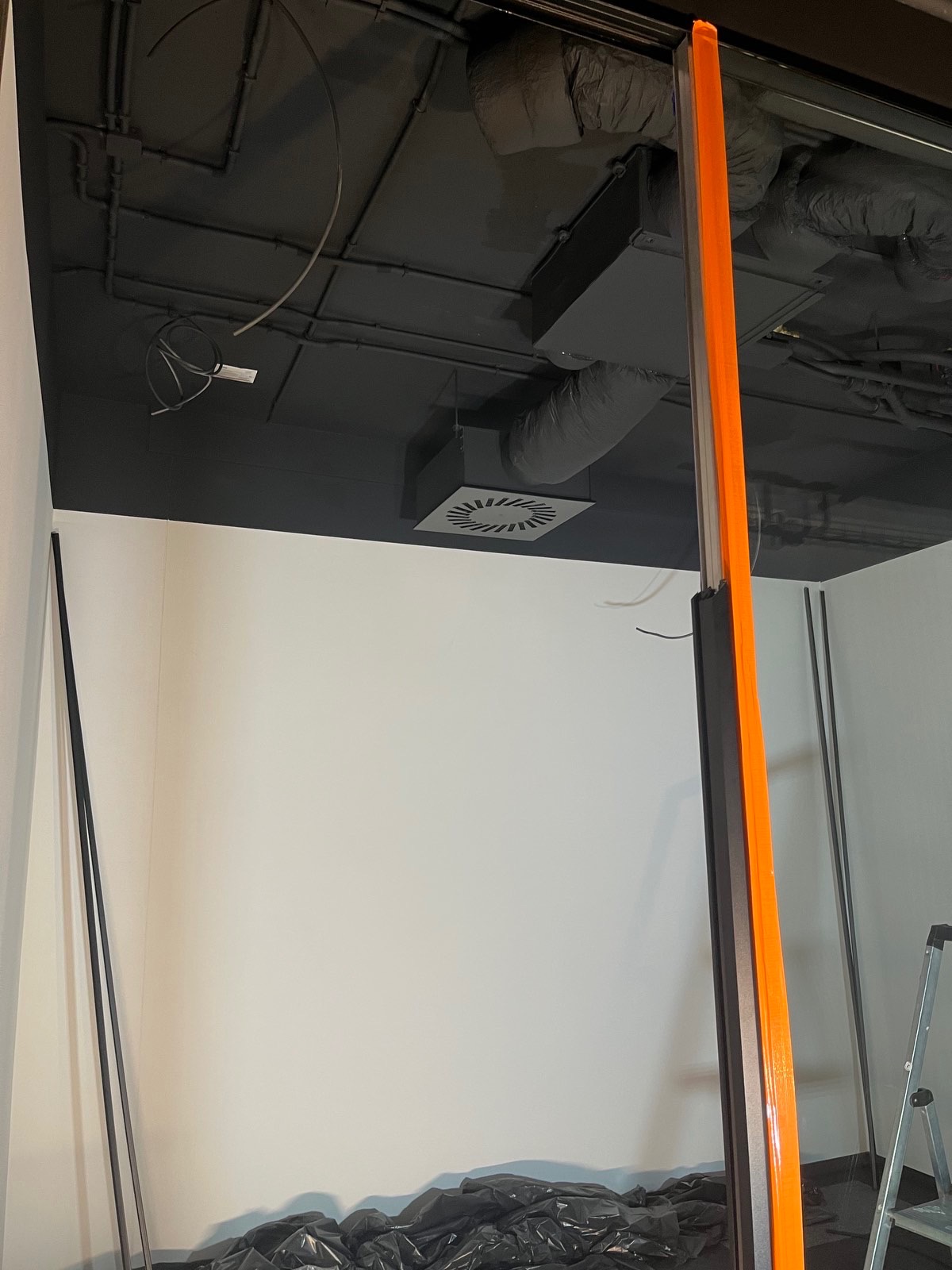 Przygotowane pomieszczenie biurowe przed pracami: czarny sufit z instalacjami, białe ściany, zabezpieczona folią podłoga, widoczna drabina malarska.