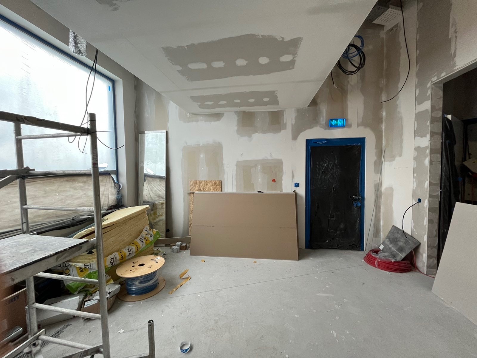 Wnętrze w trakcie prac wykończeniowych: szpachlowane ściany i sufit, płyty gipsowo-kartonowe, izolacja, rusztowanie i kable na podłodze.