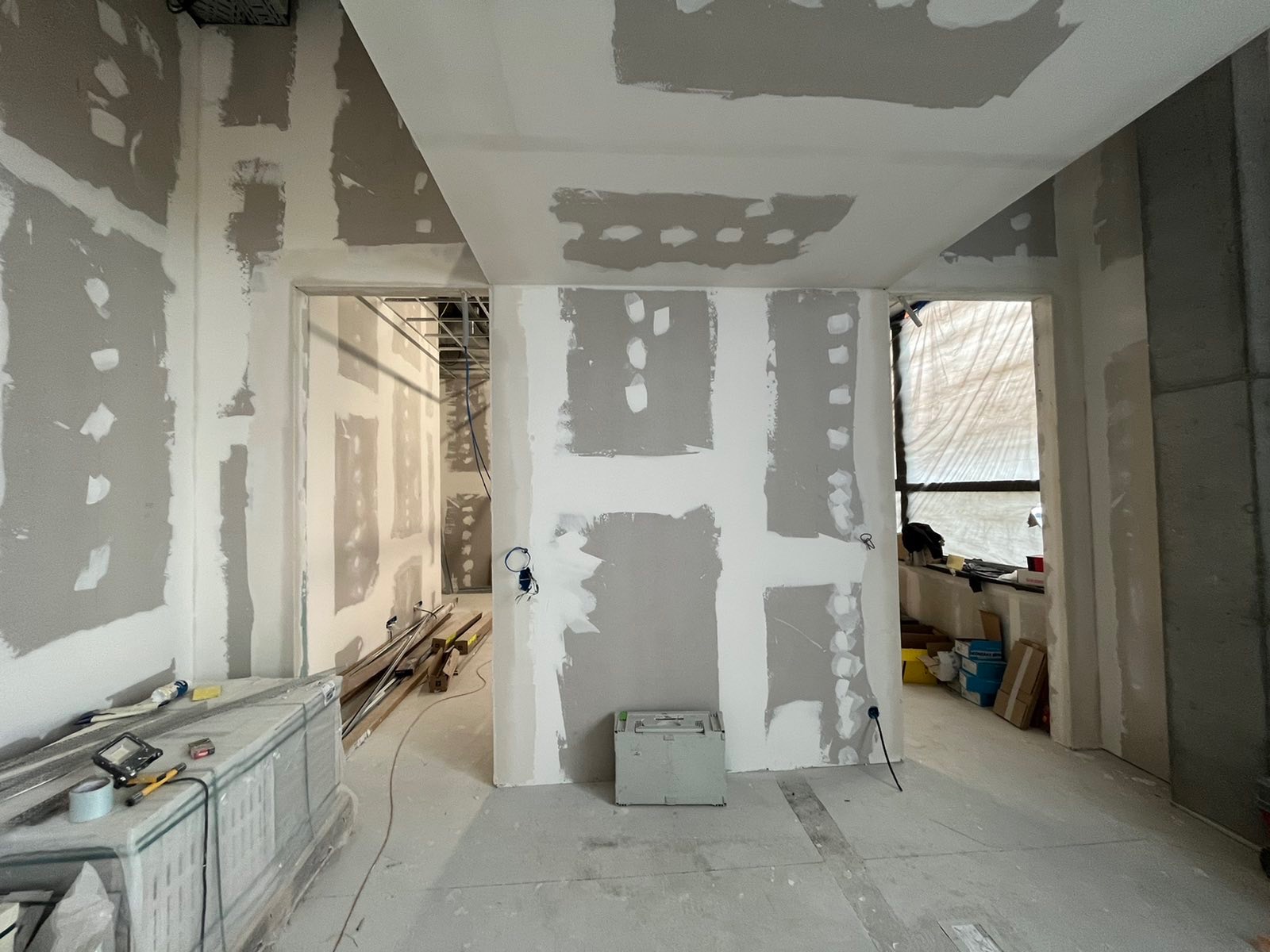 Wnętrze w trakcie remontu: ściany pokryte gładzią gipsową, widoczne przewody elektryczne, materiały budowlane i narzędzia na podłodze, przygotowanie do malowania.