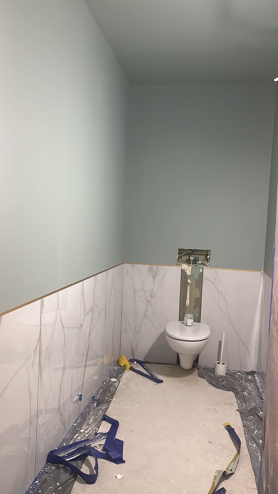 Łazienka w trakcie remontu: pomalowane na pastelowy odcień zieleni ściany, ułożone marmurowe płytki do połowy wysokości, zabezpieczona folią podłoga, widoczny fragment instalacji nad toaletą.