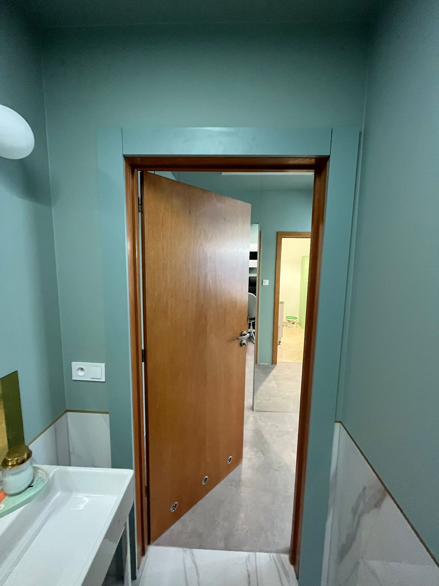 Świeżo pomalowane na turkusowo ściany łazienki z widoczną białą umywalką i otwartymi drzwiami do kolejnego pomieszczenia.