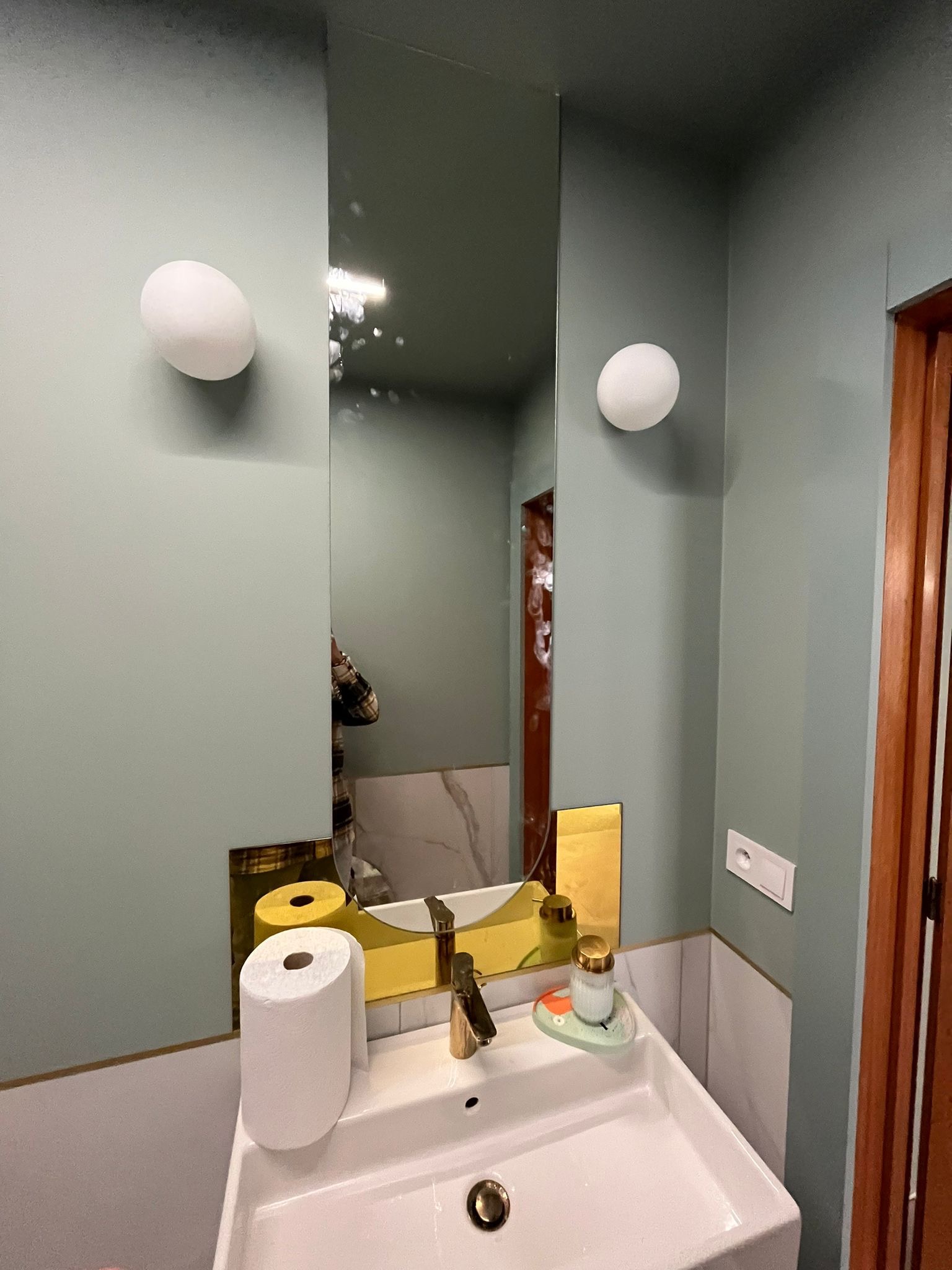 Umywalka w łazience z lustrem, okrągłymi lampami ściennymi i zielonymi ścianami, widoczne detale wykończenia i dozownik mydła.