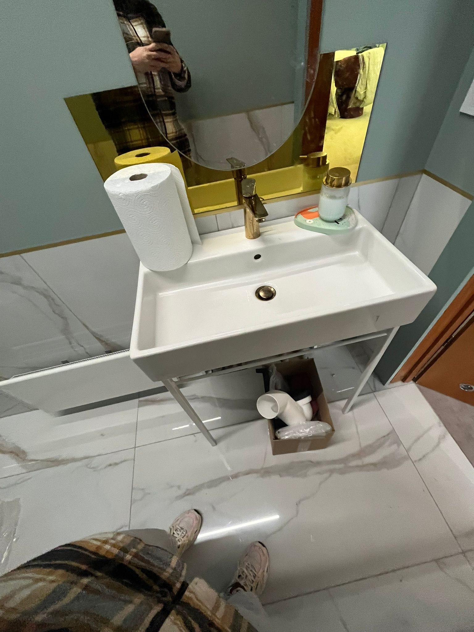 Nowa, biała umywalka na metalowym stelażu w łazience z marmurową podłogą, widoczne elementy instalacji w kartonie pod umywalką, odbicie osoby robiącej zdjęcie w lustrze.