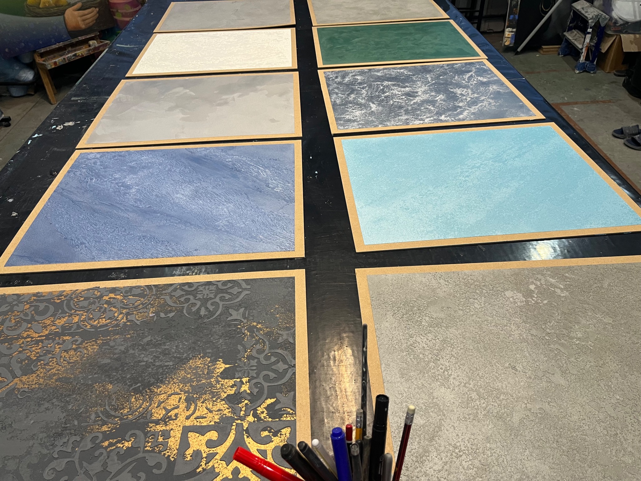 Wzornik z różnymi fakturami i kolorami tynków dekoracyjnych na drewnianych ramkach, widoczne narzędzia malarskie w pojemniku.