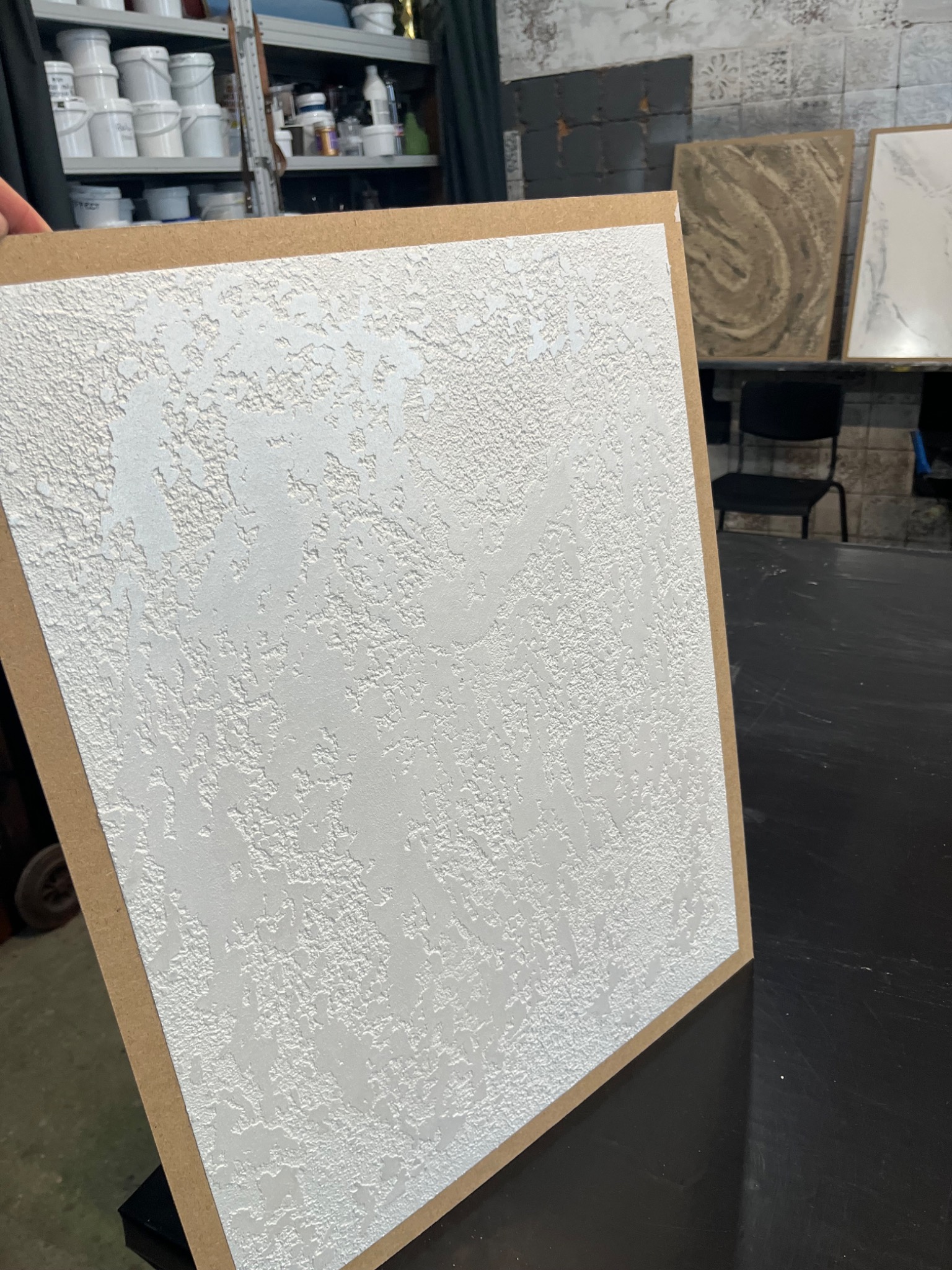 Próbka białej, strukturalnej farby na płycie, prezentująca efekt dekoracyjny z wyraźną fakturą, umieszczona w warsztacie na tle innych próbek i narzędzi.