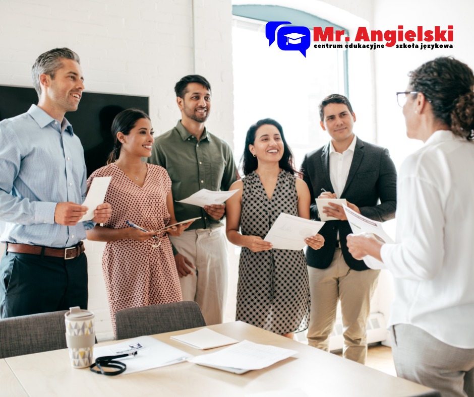 Grupa uśmiechniętych osób z dokumentami w dłoniach, uczestniczących w spotkaniu biznesowym lub zajęciach językowych w Gnieźnie. W tle widoczne logo szkoły językowej.