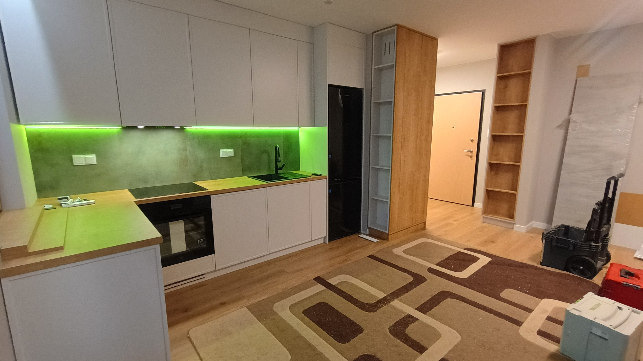 Nowoczesna kuchnia z białymi szafkami, drewnianym blatem i zielonym oświetleniem LED. W tle widoczny regał i narzędzia, sugerujące etap wykończenia.