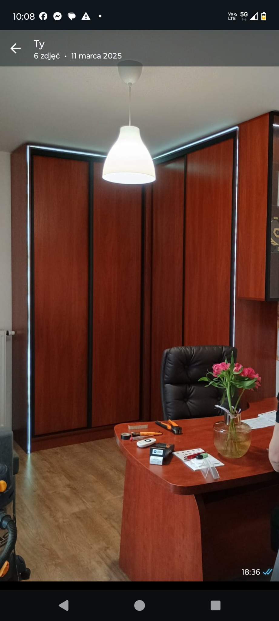 Wnętrze z szafą wnękową z oświetleniem LED, biurkiem i kwiatami. Widoczne narzędzia na blacie biurka. Ujęcie z perspektywy podłogi, podkreślające wysokość zabudowy.