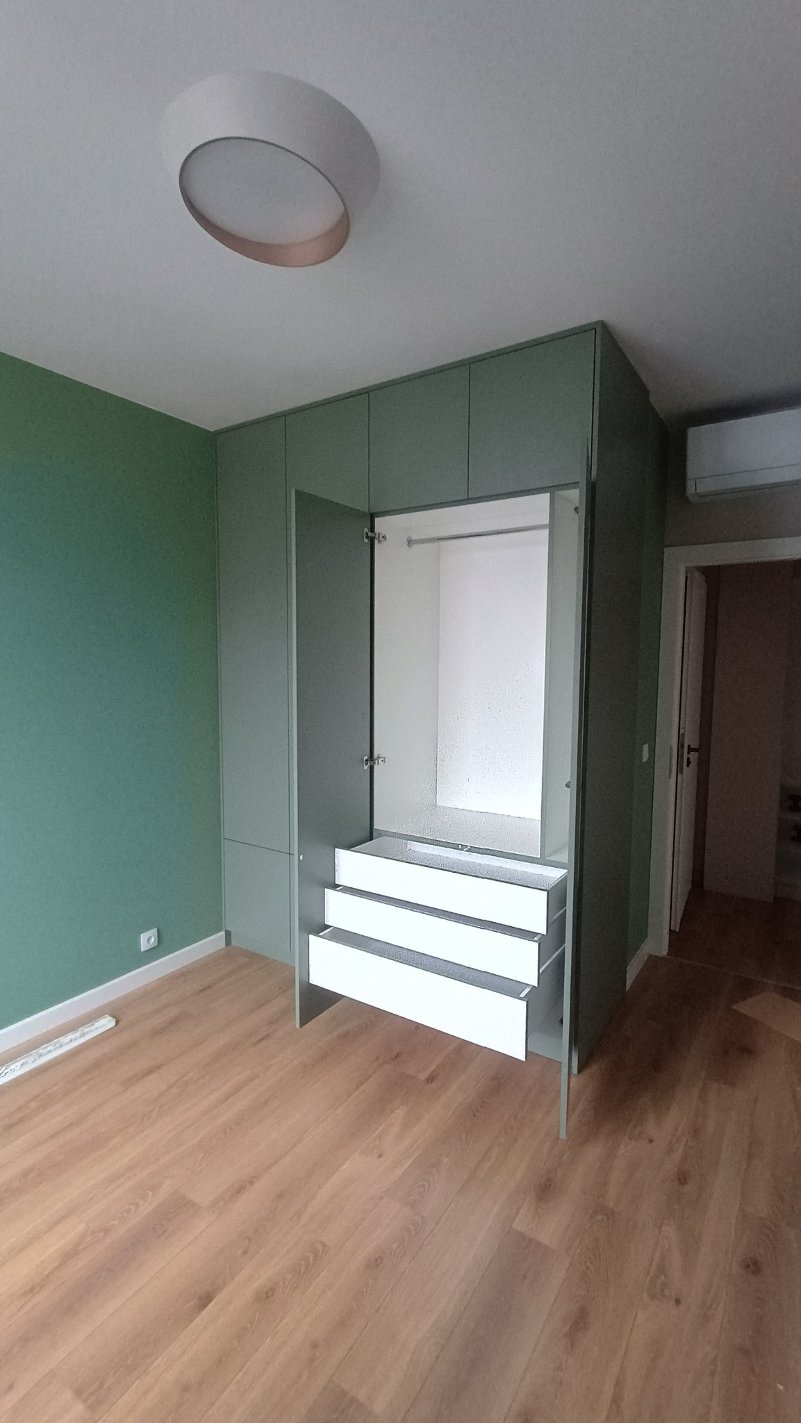 Jasnozielona szafa wnękowa z otwartymi drzwiami i wysuniętymi białymi szufladami, na tle drewnianej podłogi i zielonej ściany. Widoczny okrągły plafon.