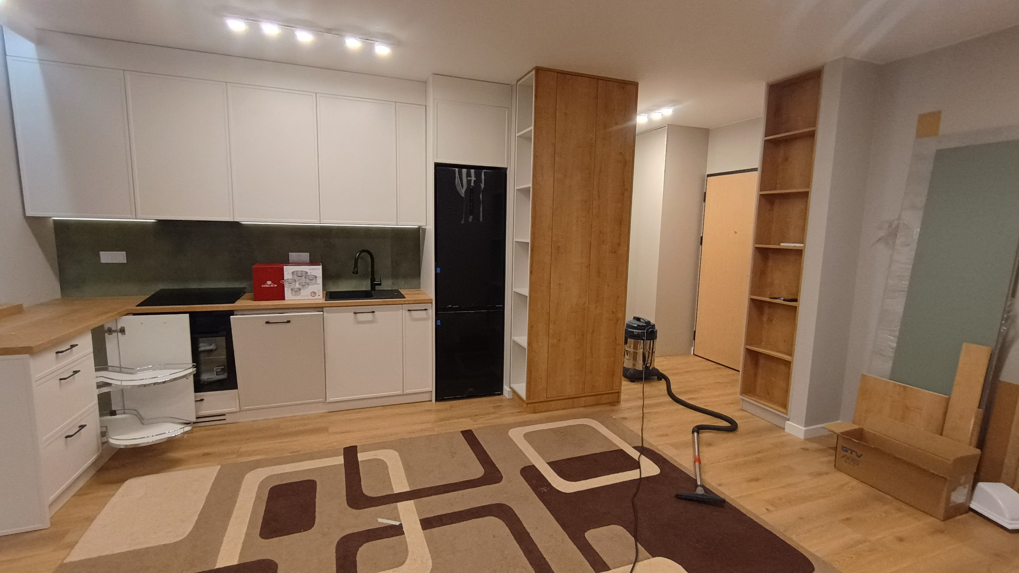 Jasna kuchnia z drewnianym blatem i czarną lodówką. Widoczny otwarty narożny kosz cargo. Na podłodze wzorzysty dywan i odkurzacz. W tle regał i materiały budowlane.