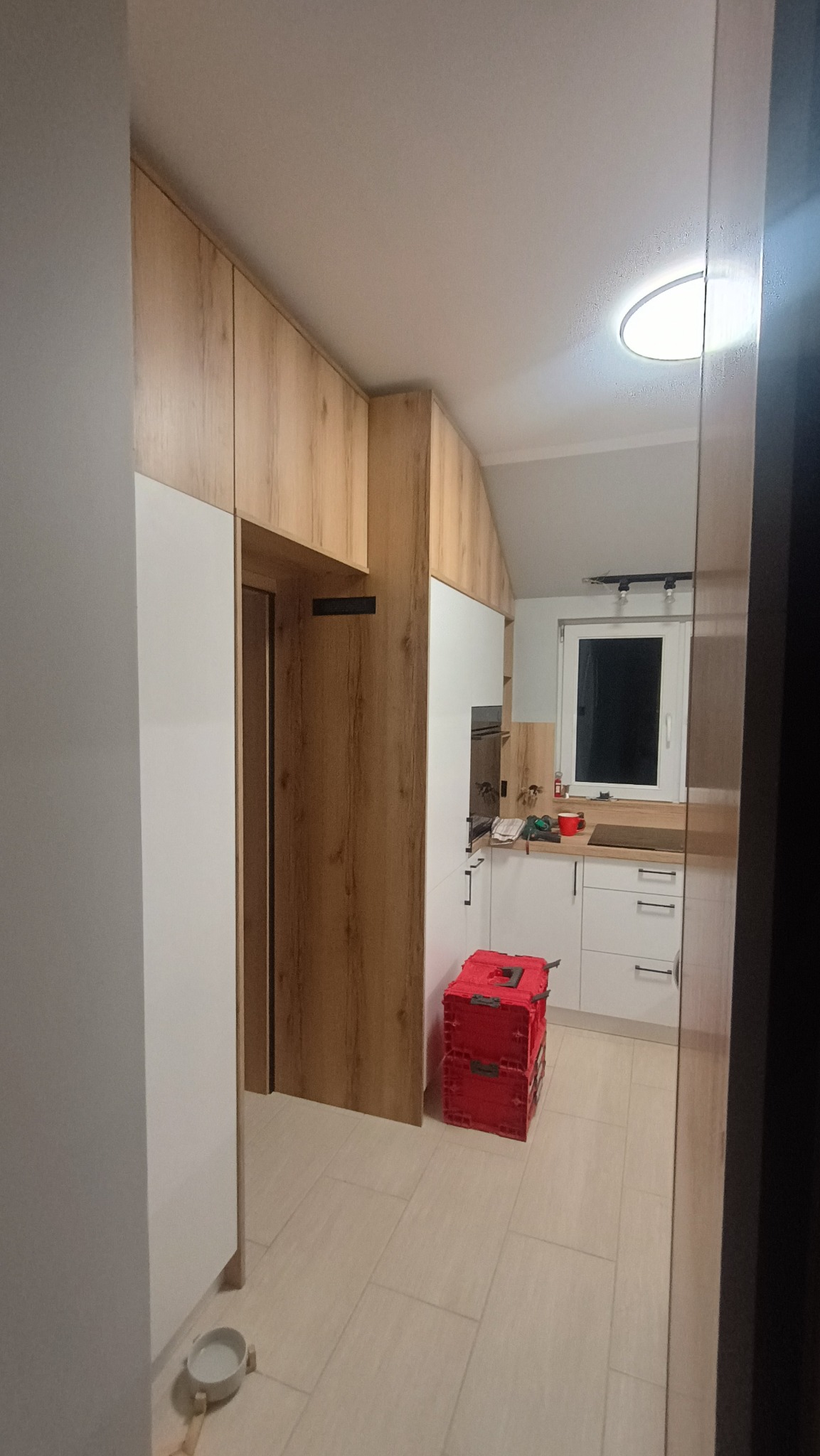 Jasna kuchnia z drewnianymi akcentami, widok z korytarza. Czerwony pojemnik na podłodze dodaje koloru. Uwagę przykuwa oświetlenie sufitowe i minimalistyczne uchwyty.