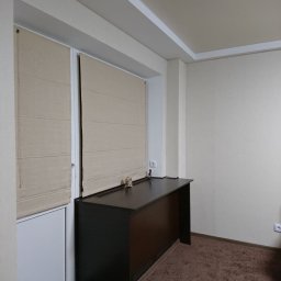 A Line interiors - Beżowe rolety rzymskie zamontowane w oknie balkonowym, z ciemnobrązowym biurkiem przylegającym do ramy okiennej, na tle beżowych ścian i brązowej wykładziny dywanowej.