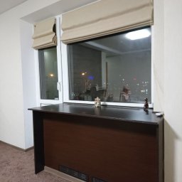 A Line interiors - Balkon z zabudową okienną i brązowym blatem, ozdobiony figurkami, z beżowymi roletami rzymskimi.