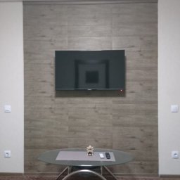 A Line interiors - Telewizor zawieszony na ścianie wyłożonej panelami imitującymi drewno, szklany stolik kawowy z dekoracją i pilotami, brązowy dywan.