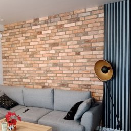 A Line interiors - Ceglana ściana z dekoracyjnymi panelami pionowymi w kolorze grafitowym, szara sofa z poduszkami i lampa stojąca z kloszem w kolorze złota.